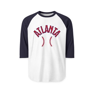 May include: White and navy blue baseball-style shirt. The word "ATLANTA" is arched across the chest in red letters with a baseball stitch design below. The shirt has navy blue sleeves.