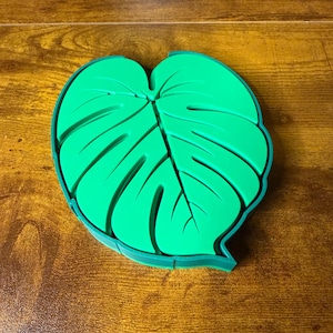 May include: A bright green, leaf-shaped coaster with a detailed vein pattern. The coaster has a dark green border and is designed to resemble a Monstera plant leaf. The coaster is made of a flexible material.