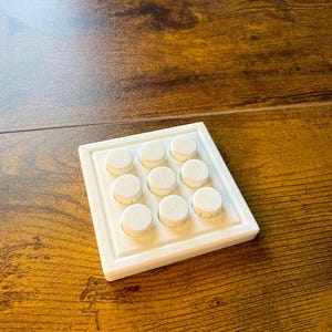 May include: A white, square jewellery display tray with nine circular, white, textured pads. The tray sits on a dark brown, wood-grain surface. The pads are evenly spaced within the tray's recessed area.