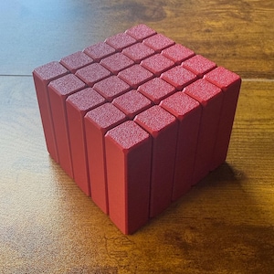 May include: A red cube puzzle composed of smaller rectangular blocks. The blocks are arranged to form a larger cube shape. The surface of the blocks has a textured appearance, and the color is a solid, vibrant red.