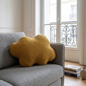 May include: A mustard-yellow cloud-shaped cushion sits on a grey sofa. The cushion has a textured, fluffy appearance. The sofa is in a room with a window and a stack of books.