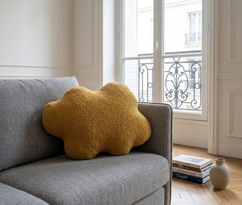 May include: A mustard-yellow cloud-shaped cushion sits on a grey sofa. The cushion has a textured, fluffy appearance. The sofa is in a room with a window and a stack of books.