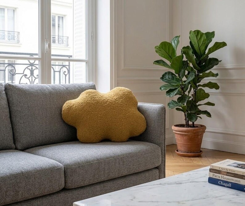 May include: A mustard-yellow cloud-shaped cushion on a grey sofa. A large potted plant with green leaves sits in a terracotta pot. The room has a window and a marble coffee table.