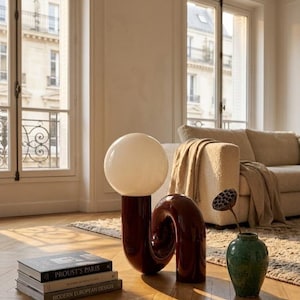 Peut inclure: Intérieur moderne avec une lampe sphérique blanche sur un socle rouge foncé, une sculpture assortie et un vase vert avec des fleurs séchées. Des livres sont empilés sur le sol près de la lampe.
