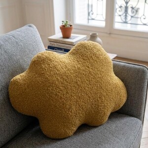 May include: Mustard yellow cloud-shaped cushion with a textured, plush surface. The cushion is placed on a grey sofa, offering a soft aesthetic. This accent cushion adds a touch of whimsy to any living space. Measures approximately 40cm across.