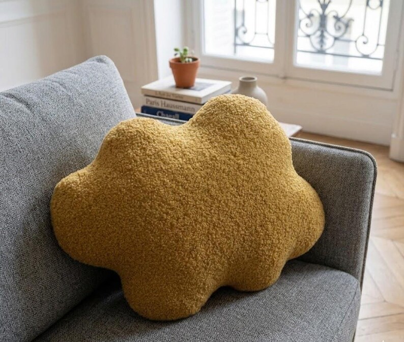 May include: Mustard yellow cloud-shaped cushion with a textured, plush surface. The cushion is placed on a grey sofa, offering a soft aesthetic. This accent cushion adds a touch of whimsy to any living space. Measures approximately 40cm across.
