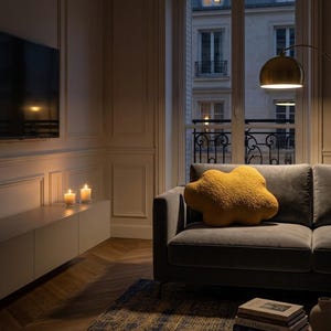 May include: A living room with a grey sofa and a mustard-yellow cloud-shaped cushion. A gold-shaded floor lamp provides light. Two lit candles sit on a white cabinet beneath a wall-mounted TV. A window offers a view of a city.
