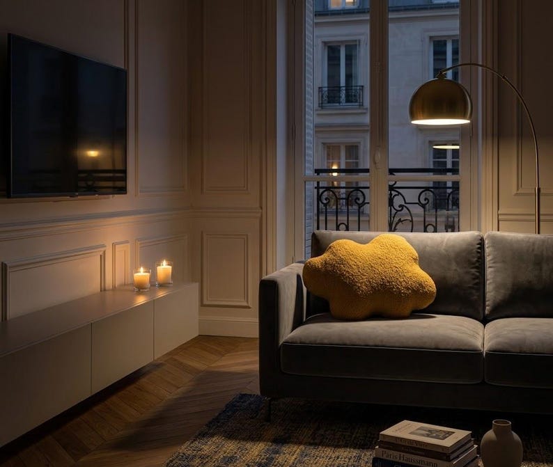 May include: A living room with a grey sofa and a mustard-yellow cloud-shaped cushion. A gold-shaded floor lamp provides light. Two lit candles sit on a white cabinet beneath a wall-mounted TV. A window offers a view of a city.