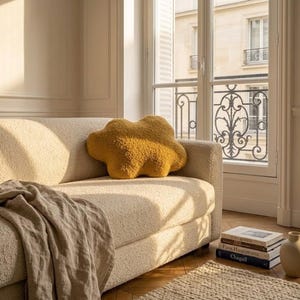 May include: A mustard-yellow cloud-shaped cushion sits on a cream sofa. A beige throw is draped over the sofa. The room has a window with a view of a building and a stack of books.