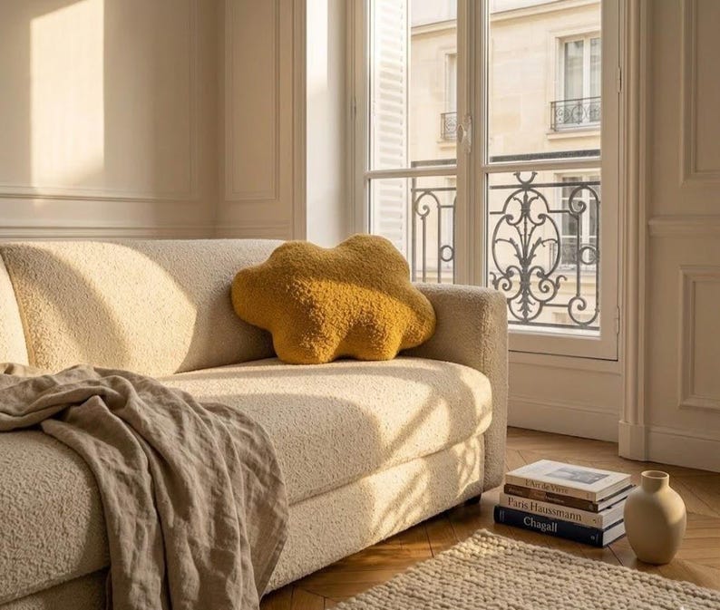 May include: A mustard-yellow cloud-shaped cushion sits on a cream sofa. A beige throw is draped over the sofa. The room has a window with a view of a building and a stack of books.