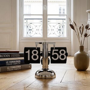 Peut inclure: Une horloge à bascule chromée affiche 18h58. Des livres et un stylo sont posés sur le parquet. Un vase avec des fleurs séchées est près d'une fenêtre. La pièce a un design classique et élégant.
