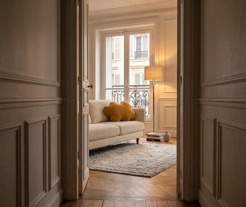 May include: Interior view of a living room with a cream sofa and a yellow cloud-shaped cushion. A patterned rug rests on a wooden floor. A floor lamp and a window with shutters are in the background.