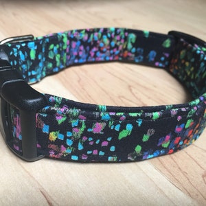 May include: A black dog collar with a colorful, abstract pattern of blue, green, pink, and yellow spots. The collar has a black plastic buckle and is designed for a pet.