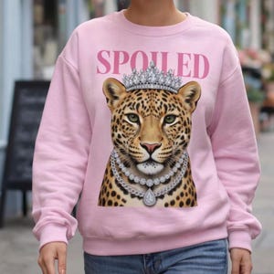 May include: A light pink crewneck sweatshirt featuring a graphic of a leopard wearing a silver crown and layered diamond necklaces. The word SPOILED is printed in pink capital letters above the leopard's head. The sweatshirt has long sleeves.