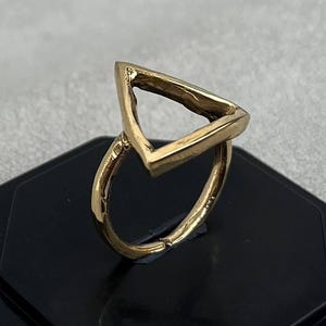 May include: A gold-toned ring featuring a prominent open triangle design. The band is thin and circular, connecting to the base of the triangular top. The metal surface has a polished finish with visible texture along the edges of the geometric shape.