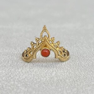 May include: A gold-toned metal ring featuring an intricate, pointed crown-like design with a small, round, orange-red stone set in the center. The band has a decorative, filigree-style pattern with small bead-like details along the edges.