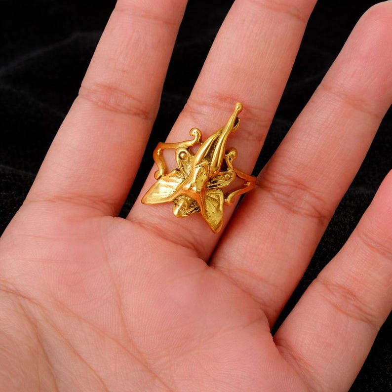 May include: A gold-colored ring featuring a floral design with a central figure. The ring has an ornate, detailed appearance, with a textured surface. The ring is set on a finger, showcasing its size and design.