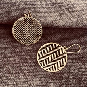 Puede incluir: Dos pendientes circulares plateados con un patrón en zigzag. Los pendientes tienen un diseño calado y un gancho. Se muestran sobre una superficie texturizada de color gris oscuro.