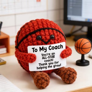 May include: A handmade, red crocheted basketball figure with black accents and a smiling face, holding a white sign that reads "To My Coach." A small basketball on a stand is also visible.