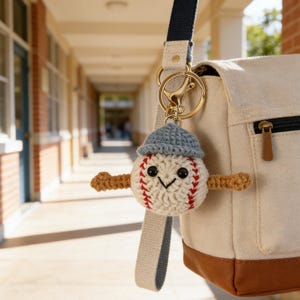 Puede incluir: Un llavero con temática de béisbol con un personaje de béisbol de ganchillo que lleva una gorra azul y brazos marrones. El llavero está sujeto a una bolsa beige con una base marrón y una correa azul marino. El fondo muestra un pasillo escolar.