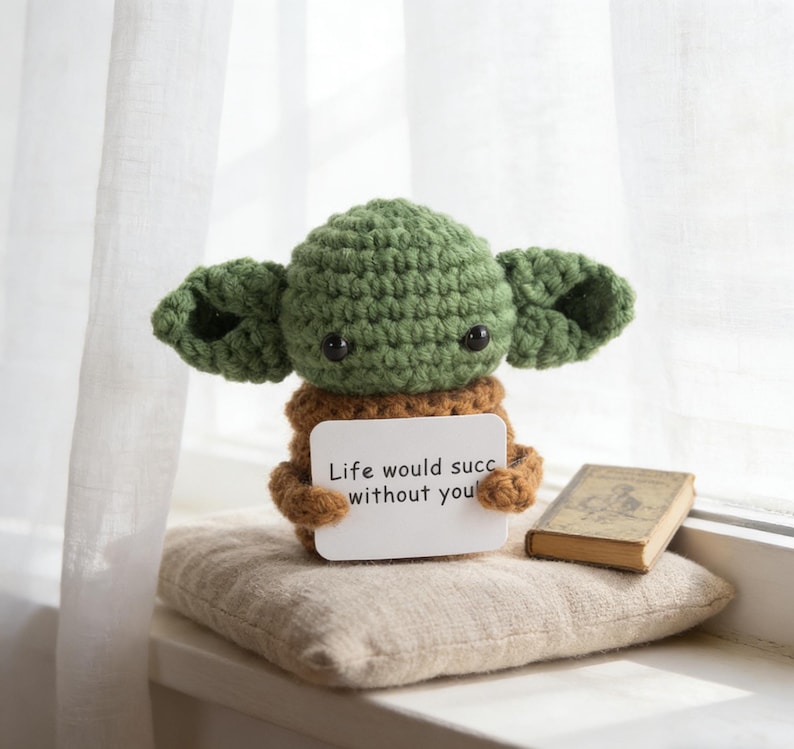 K&ouml;nnte beinhalten: Eine geh&auml;kelte gr&uuml;ne und braune Figur mit gro&szlig;en Ohren, die ein Schild mit der Aufschrift "Life would succ without you" h&auml;lt. Die Figur sitzt auf einem kleinen Kissen neben einem Miniaturbuch.
