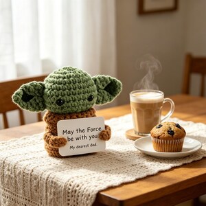 May include: A crocheted green and brown figure resembling a character from a popular science fiction franchise, holding a card with the text "May the Force be with you. My dearest dad." A cup of coffee and a muffin are on the table.