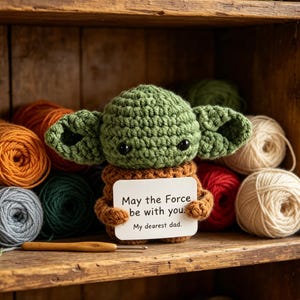 May include: A crocheted green character with large ears, holding a sign that reads "May the Force be with you. My dearest dad." The character is brown and green, with black eyes. The background includes yarn in various colors.