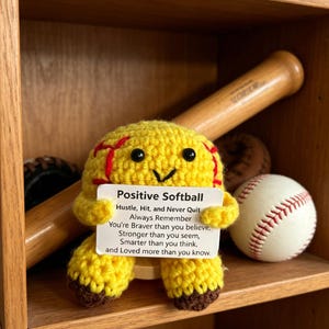 Puede incluir: Una figura de softbol amarilla tejida a ganchillo con una cara sonriente y costuras rojas, sosteniendo un letrero que dice "Positive Softball". Un bate de béisbol de madera y varias pelotas de béisbol están en el fondo, exhibidos en un estante de madera.
