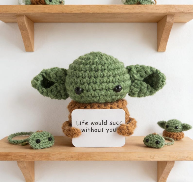 K&ouml;nnte beinhalten: Geh&auml;kelte gr&uuml;ne Figur mit gro&szlig;en Ohren, die ein Schild mit der Aufschrift "Life would succ without you!" h&auml;lt. Mehrere andere geh&auml;kelte Figuren befinden sich auf einem Holzregal. Die Figuren sind braun und gr&uuml;n.