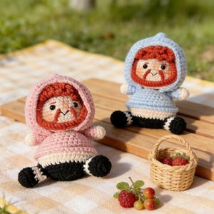 May include: Two handmade crochet dolls, one in pink and the other in blue, sit on a wooden surface. Each doll has a hooded head, a red beard, and black and white striped legs. A small woven basket filled with berries and some loose berries are also in the image.