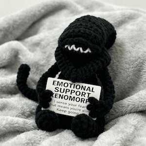 Könnte beinhalten: Eine schwarze, gehäkelte Xenomorph-Figur mit weißen Zähnen und einem strukturierten Körper. Sie hält eine weiße Karte mit der Aufschrift "EMOTIONAL SUPPORT XENOMORPH" und ermutigendem Text. Die Figur befindet sich vor einem weichen, grauen Hintergrund.
