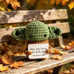 May include: A green crocheted figure with large ears, holding a white card with the text "May the Force be with you. My dearest dad." The figure is sitting on a wooden bench with fall leaves.