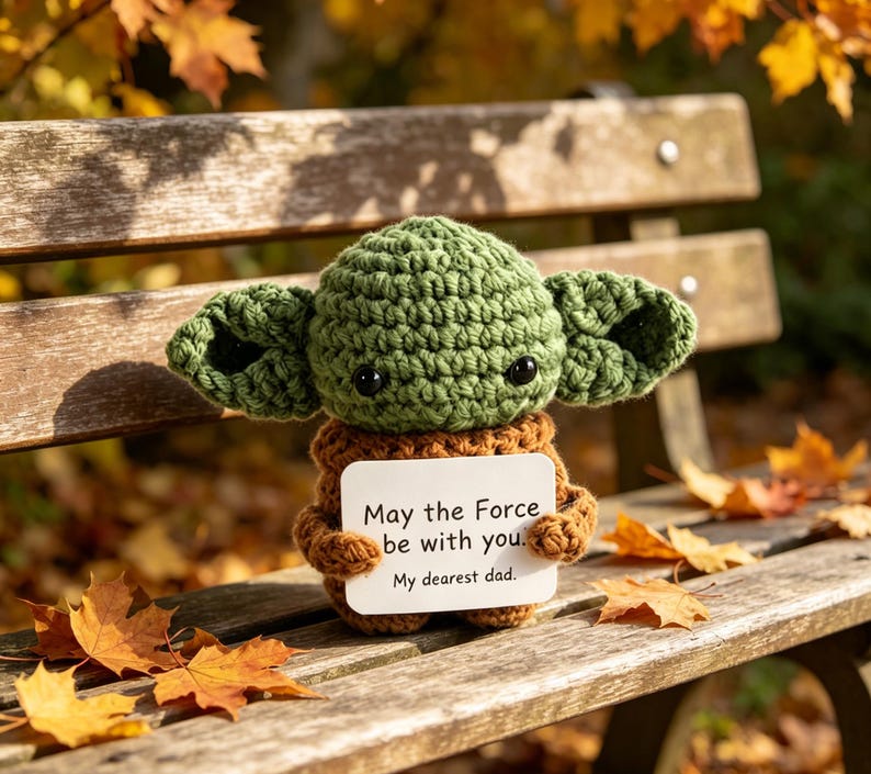 May include: A green crocheted figure with large ears, holding a white card with the text "May the Force be with you. My dearest dad." The figure is sitting on a wooden bench with fall leaves.