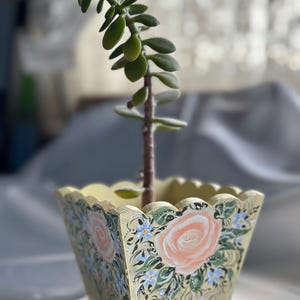 Op de afbeelding: Een kleine vetplant in een handbeschilderde, vierkante houten plantenbak. De plantenbak is lichtgeel geschilderd met een geschulpte bovenrand en heeft een bloemenontwerp met perzikrozen en blauwe accenten.