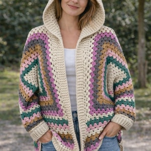 May include: A hooded, open-front crochet cardigan in a cream color with a colorful geometric pattern. The pattern includes shades of pink, green, gray, and tan. The cardigan is worn with blue jeans.