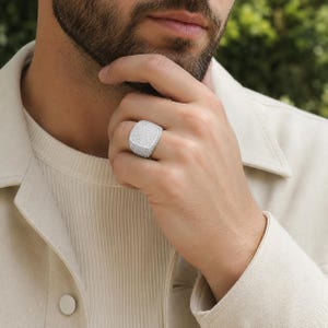 May include: A square-shaped signet ring featuring a pavé setting with numerous small, clear, reflective stones covering the entire top surface and the sides of the band, creating a bright, metallic, and highly textured appearance on the finger.