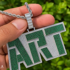 May include: A silver-toned pendant necklace featuring the word ART in block letters. The pendant is encrusted with small green stones on the face and clear stones along the borders and the bail, attached to a silver-toned rope chain.