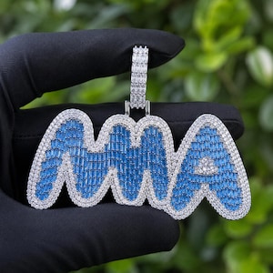 May include: A pendant spelling "MA" in a blue and white design. The letters are outlined in white stones, with blue stones filling the interior. The pendant hangs from a silver-colored bail.