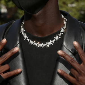 May include: A silver-coloured necklace featuring a barbed wire design. The necklace is worn around the neck and is made of shiny metal. The person in the image is wearing a black shirt and a black leather jacket.