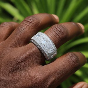 May include: A wide, silver-toned ring encrusted with numerous small, round, and rectangular-cut clear stones. The ring is on a person's finger, with a green leafy background.