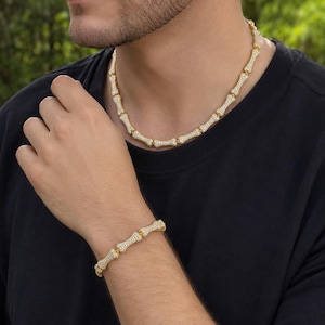 May include: A gold-toned chain necklace and a matching bracelet, both featuring a series of connected bone-shaped links. Each link is encrusted with numerous small, clear, sparkling stones that cover the entire surface of the gold-colored metal.