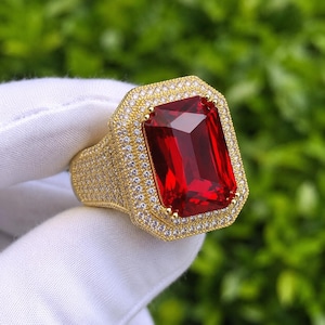 Puede incluir: Un anillo dorado con una gran piedra preciosa roja oscura de forma rectangular. El anillo está engastado con pequeñas piedras preciosas claras alrededor de la piedra central y en la banda, creando un efecto brillante. El anillo se sostiene sobre un fondo verde.