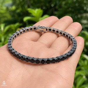 Könnte beinhalten: Ein Tennisarmband mit einer durchgehenden Reihe runder, facettierter schwarzer Steine, die in einen polierten, silberfarbenen Metallrahmen gefasst sind. Das Armband verfügt über einen sicheren Kastenschloss-Verschluss und eine flexible Gliederkonstruktion.