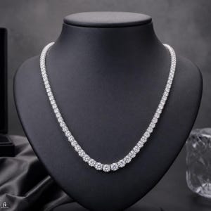 Peut inclure: Un collier tennis argenté composé d'une rangée continue de pierres rondes, transparentes et facettées, serties dans une monture métallique. Le collier est présenté sur un buste noir, mettant en valeur la régularité des pierres scintillantes.