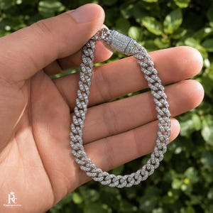 May include: A silver-toned cuban link bracelet featuring a continuous pattern of small, clear, sparkling stones embedded along each link. The bracelet includes a rectangular box clasp closure that is also encrusted with clear stones.
