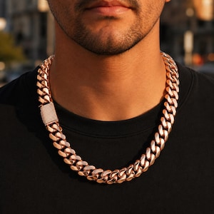 Puede incluir: Un collar de cadena de eslabones cubanos grueso en tono oro rosa con un cierre de caja rectangular incrustado con numerosas piedras pequeñas, claras y reflectantes. Los eslabones metálicos están pulidos y tienen una superficie lisa.