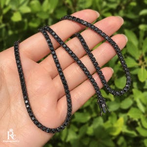 May include: A black tennis necklace featuring a continuous row of round, dark-toned stones set in a matching black metal chain. The necklace includes a secure box clasp closure and is displayed in a coiled arrangement to show the link structure.