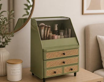 以下が含まれることがあります： オリーブグリーンのデスクトップオーガナイザー。ブラシや化粧品用の透明な上部コンパートメント、幅広の引き出し2つ、下部の小さな引き出し2つを備え、編み込みラタンパネルと黒いノブで装飾され、短い黒い脚が付いています。