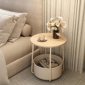 Op de afbeelding: Een kleine, ronde bijzettafel met een licht houten blad en witte metalen poten. Op de tafel staat een kopje koffie naast een vaas met bloemen. Een geweven mand eronder bevat boeken en een bril.