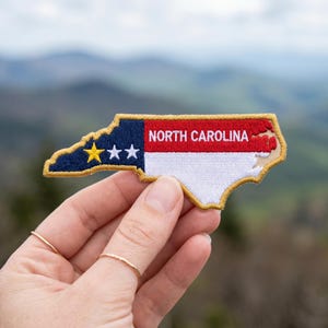 May include: An embroidered patch shaped like the state of North Carolina featuring the state flag design. The patch has a blue section with one yellow star and two white stars, followed by red and white horizontal stripes with the text NORTH CAROLINA.